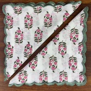 May include: Two white floral napkins with pink flower and green leaf patterns. The napkins have a scalloped green border. One napkin is folded in half, creating a triangle shape.