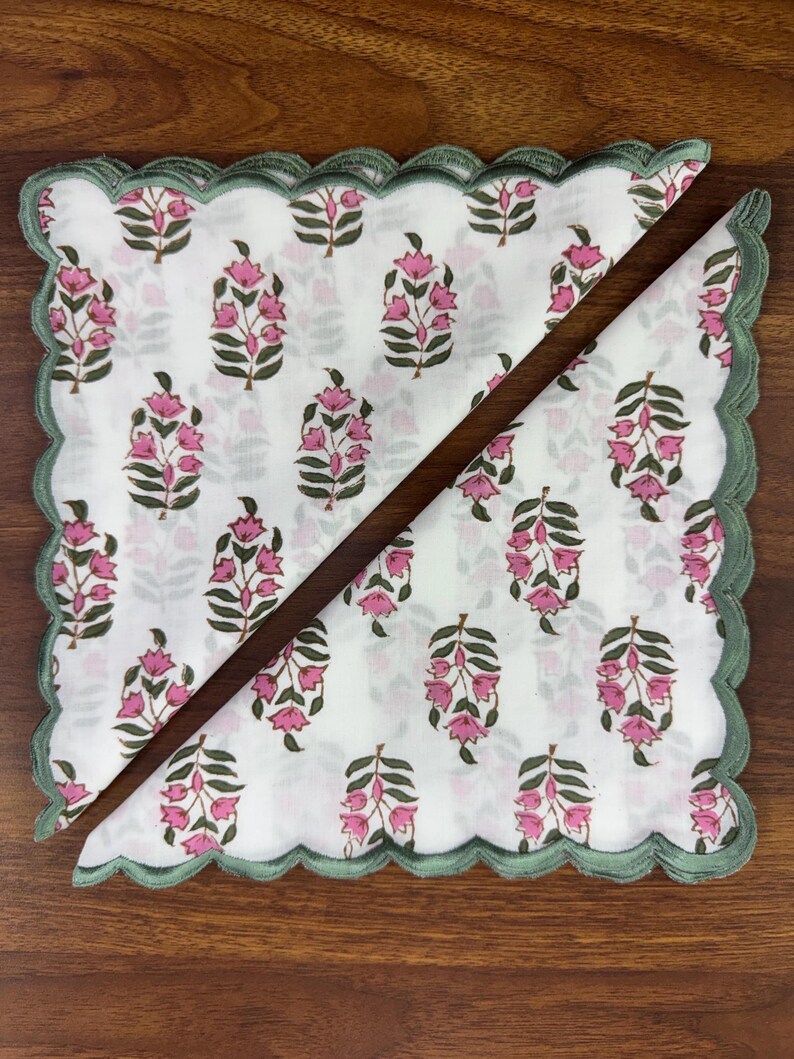 May include: Two white floral napkins with pink flower and green leaf patterns. The napkins have a scalloped green border. One napkin is folded in half, creating a triangle shape.