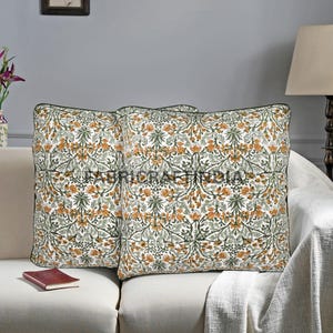 May include: Two square throw pillows with a floral pattern in orange, green, and white. The pillows have a dark green border and are placed on a cream-colored sofa. A small book rests on the sofa.
