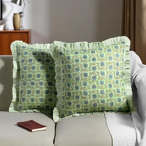 May include: Two square cushions with a light green floral pattern and frilled edges. The pattern features blue and brown floral elements on a light green background. The cushions are on a light-coloured sofa.