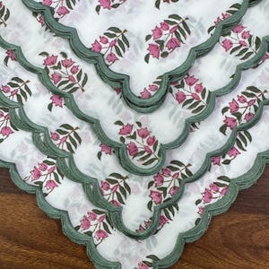 May include: White fabric napkins with a floral pattern in pink and green. The napkins have a scalloped edge in a muted green color. The floral design features pink flowers and green leaves. The napkins are arranged in a layered display.