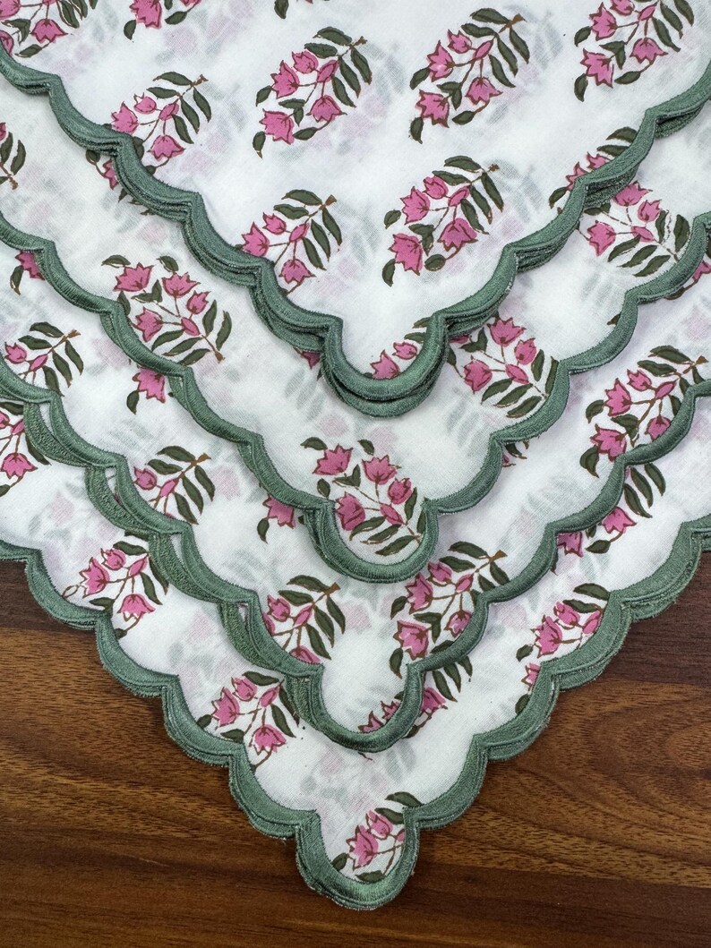 May include: White fabric napkins with a floral pattern in pink and green. The napkins have a scalloped edge in a muted green color. The floral design features pink flowers and green leaves. The napkins are arranged in a layered display.