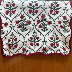 May include: A white table runner with a red and green floral pattern. The runner has a scalloped edge and a burgundy trim.