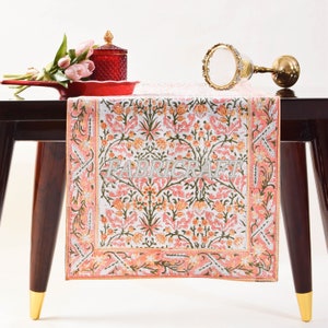 May include: A pink and white floral table runner with a green border. The runner is on a wooden table with a red tray and a glass jar with a gold lid.
