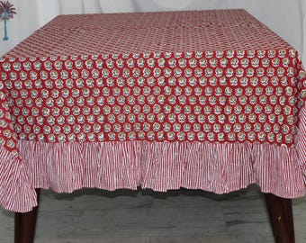 May include: A red and white patterned tablecloth with a ruffled edge. The top of the tablecloth has a repeating floral design. The ruffled edge has a red and white striped pattern. The tablecloth is on a wooden table.