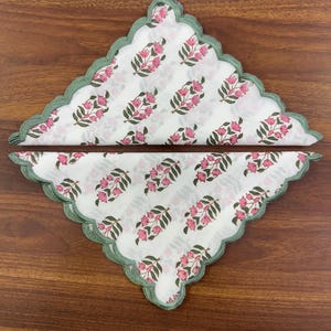May include: Two white cloth napkins with a floral pattern of pink flowers and green leaves. The napkins have a scalloped edge with a green trim. The napkins are folded and arranged on a wooden surface.
