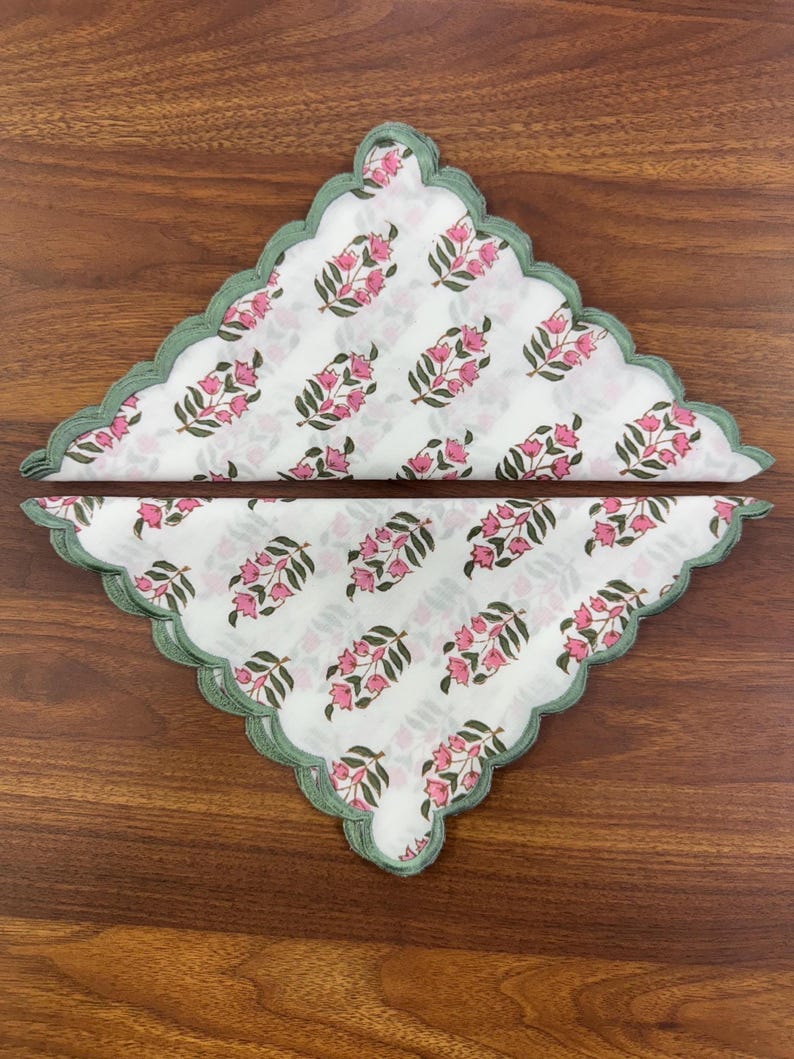 May include: Two white cloth napkins with a floral pattern of pink flowers and green leaves. The napkins have a scalloped edge with a green trim. The napkins are folded and arranged on a wooden surface.