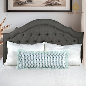 May include: A gray tufted headboard with a decorative bolster pillow and two white pillows on a bed. The bolster pillow has a blue and white floral pattern with a ruffled edge. The bed is covered with a white sheet.