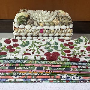 Puede incluir: Una caja decorativa cubierta de conchas marinas, con una pila de servilletas de tela dobladas en varios colores y patrones. Las servilletas presentan un dise&ntilde;o floral con bayas rojas y hojas verdes. La caja es rectangular y tiene una base de madera.