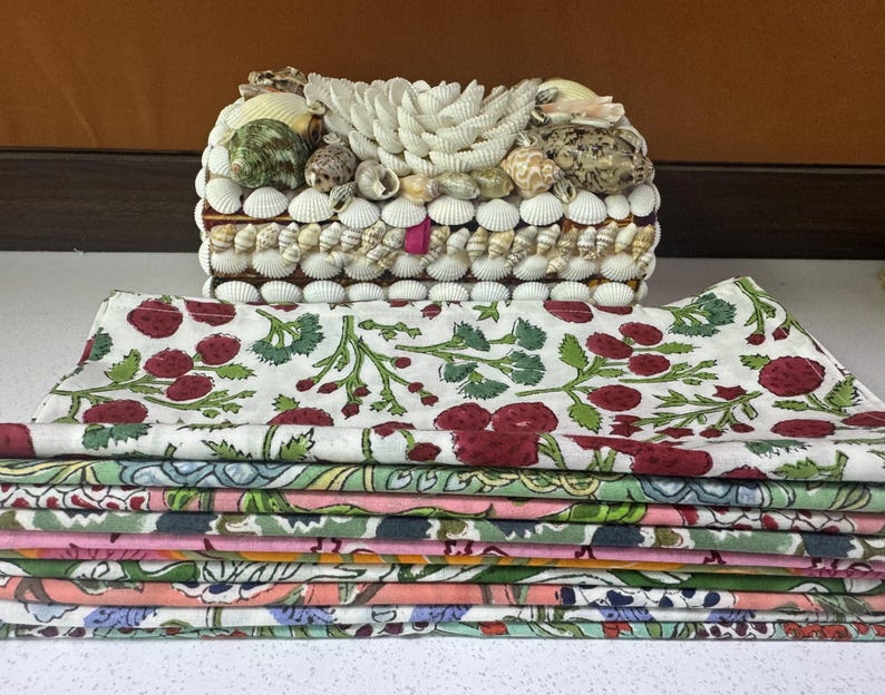 Puede incluir: Una caja decorativa cubierta de conchas marinas, con una pila de servilletas de tela dobladas en varios colores y patrones. Las servilletas presentan un dise&ntilde;o floral con bayas rojas y hojas verdes. La caja es rectangular y tiene una base de madera.