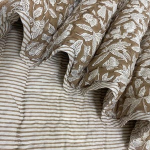 May include: A folded brown and white floral patterned quilt with a striped pattern on the bottom. The quilt features a repeating floral design in white on a brown background. The edges are trimmed with a striped border.
