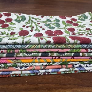 Puede incluir: Una pila de servilletas de tela coloridas con estampados florales. La servilleta superior presenta un fondo blanco con dise&ntilde;os de bayas rojas y hojas verdes. Otras servilletas muestran varios estampados florales y geom&eacute;tricos en tonos de rosa, verde y naranja.