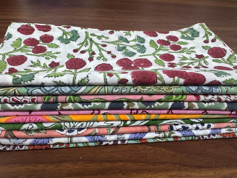 Puede incluir: Una pila de servilletas de tela coloridas con estampados florales. La servilleta superior presenta un fondo blanco con dise&ntilde;os de bayas rojas y hojas verdes. Otras servilletas muestran varios estampados florales y geom&eacute;tricos en tonos de rosa, verde y naranja.