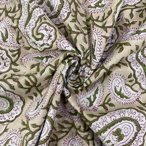 May include: Close-up of beige fabric with a repeating paisley pattern. The design features white, green, and burgundy floral motifs and swirling leaf patterns. The fabric is folded, showing the texture and detail of the print.