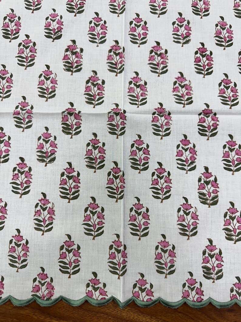 May include: White fabric with a repeating pattern of pink floral bouquets and green leaves. The fabric has a scalloped edge in a light green color. The floral design is a block print style.