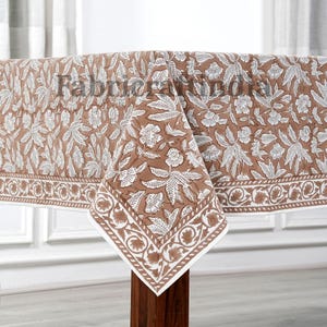 May include: A brown and white floral tablecloth with a detailed border. The tablecloth is draped over a wooden table, showcasing the intricate leaf and flower pattern. The fabric has a natural, earthy tone.