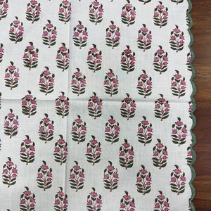 May include: White floral patterned fabric with a scalloped green border. The fabric features a repeating pattern of pink flowers with green leaves. The border is a solid green color. The fabric is likely a tablecloth or a decorative textile.