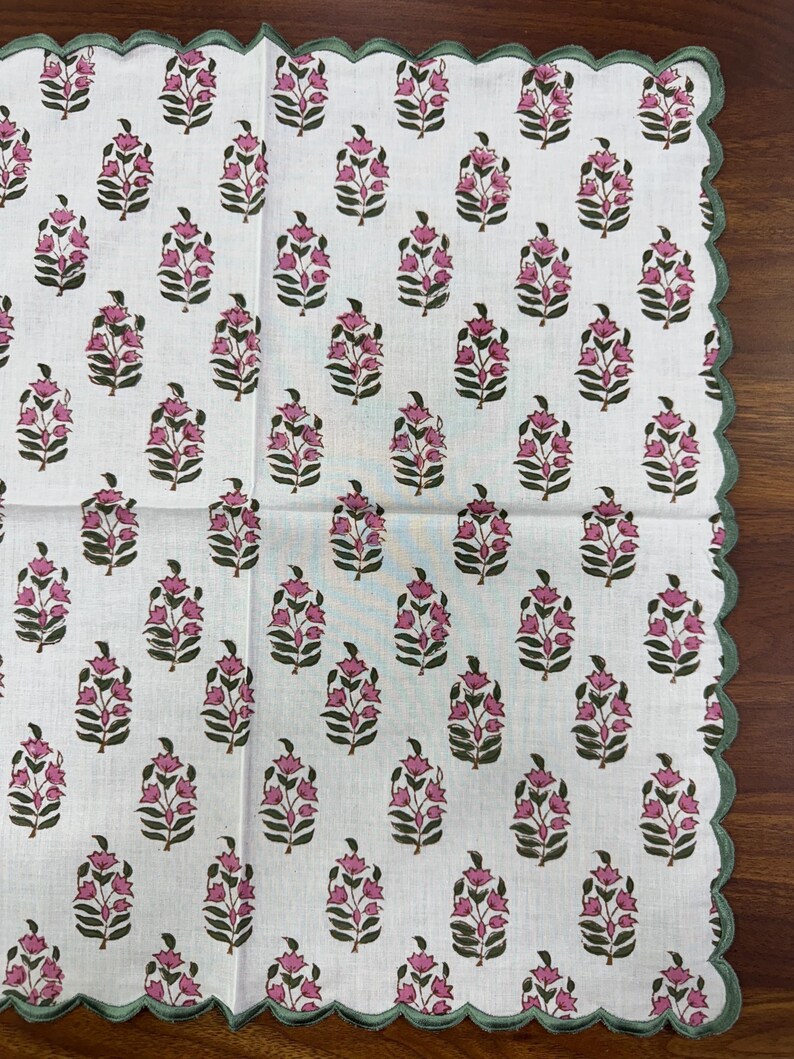 May include: White floral patterned fabric with a scalloped green border. The fabric features a repeating pattern of pink flowers with green leaves. The border is a solid green color. The fabric is likely a tablecloth or a decorative textile.