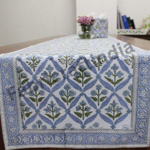 May include: A blue and white table runner with a floral pattern. The runner has a white background with blue and green floral designs. The runner has a blue border with a white floral design.