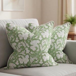 May include: Two decorative throw pillows with a green and white floral pattern rest on a gray sofa. The pillows have a soft, textured appearance and a light-colored trim. The floral design features stylized leaves and vines.