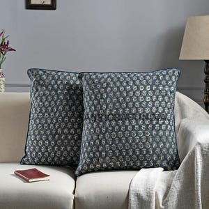 May include: Two square throw pillows with a dark blue background and a repeating floral pattern in beige and white. The pillows have a dark blue border and are placed on a beige sofa. A small book rests on the sofa.
