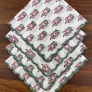 May include: A stack of white cotton napkins with a pink floral pattern and green scalloped edges. The napkins are arranged in a diamond shape, showcasing the repeating design of pink flowers and green leaves.