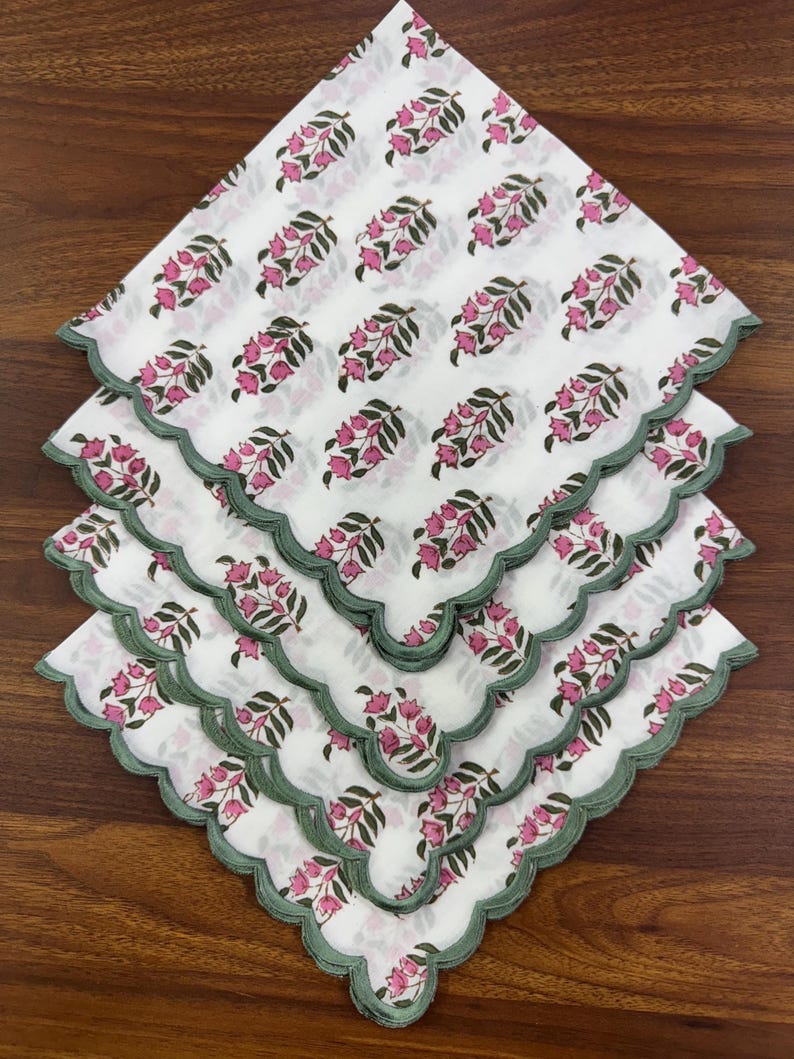 May include: A stack of white cotton napkins with a pink floral pattern and green scalloped edges. The napkins are arranged in a diamond shape, showcasing the repeating design of pink flowers and green leaves.