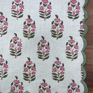 May include: White fabric with a repeating pattern of pink floral designs and green leaves. The edge is scalloped and trimmed with green stitching. The design is reminiscent of a floral block print.