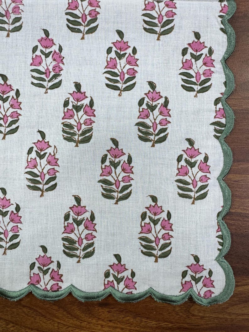 May include: White fabric with a repeating pattern of pink floral designs and green leaves. The edge is scalloped and trimmed with green stitching. The design is reminiscent of a floral block print.