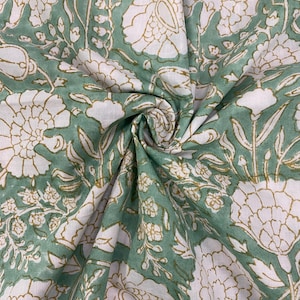 May include: Close-up of a fabric with a floral pattern. The fabric features large white flowers with gold outlines and green leaves and stems on a white background. The fabric is folded and draped.