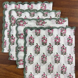 May include: Four white cloth napkins with a floral pattern of pink flowers and green leaves. Each napkin has a scalloped edge with a green border. The napkins are arranged on a wooden surface.