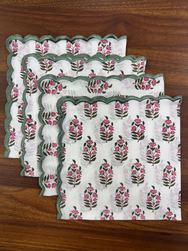 May include: Four white cloth napkins with a floral pattern of pink flowers and green leaves. Each napkin has a scalloped edge with a green border. The napkins are arranged on a wooden surface.
