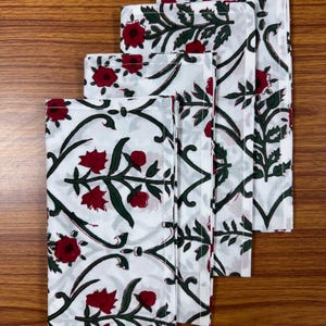 May include: Set of four white cotton napkins with a red and green floral pattern. The napkins are folded and stacked on a wooden surface.