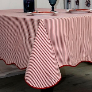 May include: A round tablecloth with a red and white striped pattern. The tablecloth has a scalloped edge with a red border. The table is set with plates, forks, and a blue glass. A vase of pink flowers is also on the table.