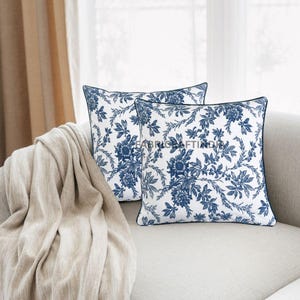 May include: Two square throw pillows with a white background and a blue floral pattern. The pillows have a dark blue trim and are placed on a light-colored sofa. A beige blanket is draped over the arm of the sofa.