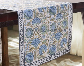 May include: A floral table runner with a white background and a repeating pattern of blue and green flowers. The border is a dark blue with a repeating geometric design. The text "FABRIC CRAFT INDIA" is printed on the runner.