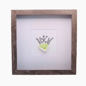 May include: A framed piece of art featuring a green sea glass piece with a black and white drawing of flowers and leaves.
