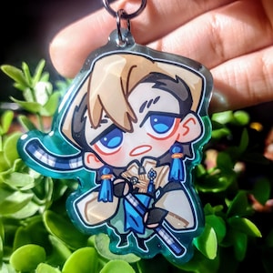 Op de afbeelding: Een heldere acryl sleutelhanger met een cartoonfiguur met blauwe ogen, blond haar en blauwe en gouden kleding. De sleutelhanger heeft een zilveren sluiting en een turquoise rand. Het personage houdt een zwaard vast.