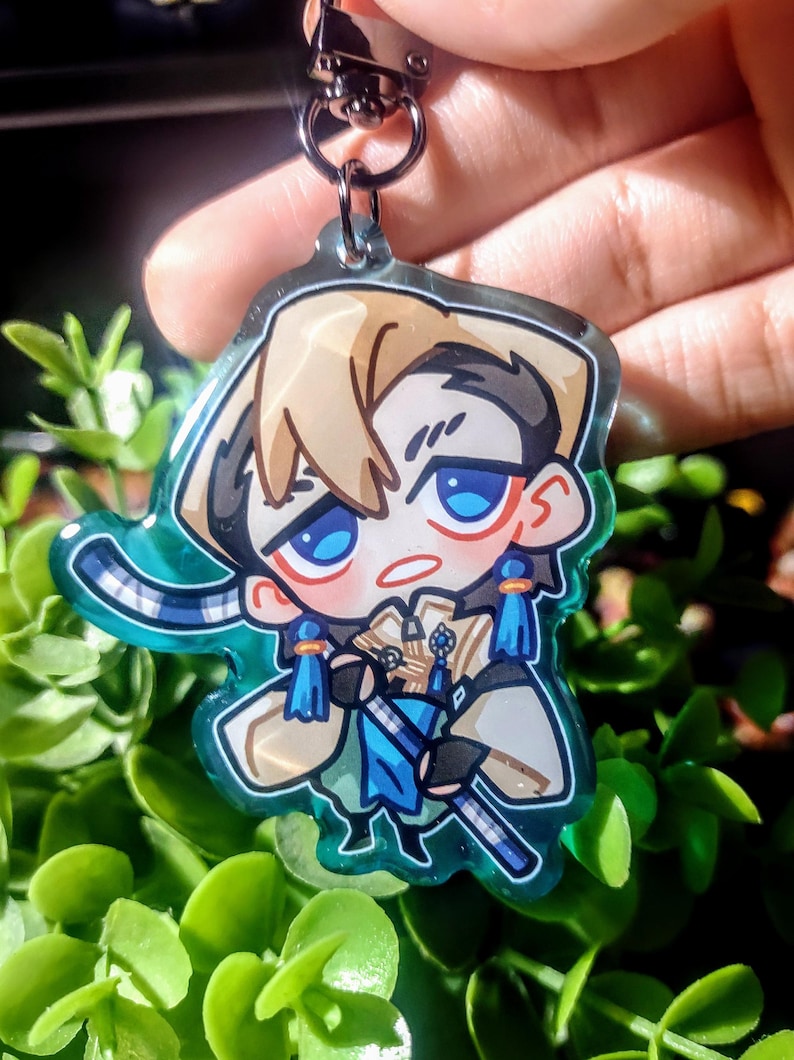 Op de afbeelding: Een heldere acryl sleutelhanger met een cartoonfiguur met blauwe ogen, blond haar en blauwe en gouden kleding. De sleutelhanger heeft een zilveren sluiting en een turquoise rand. Het personage houdt een zwaard vast.