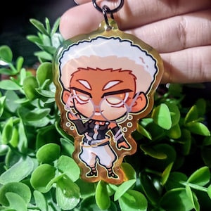 Op de afbeelding: Een heldere sleutelhanger in cartoonstijl met een personage met wit haar, een bril en een zwart-witte outfit. De sleutelhanger heeft een gouden rand en een zwarte sluiting. Het personage staat in een dynamische pose, met een bubbeleffect.