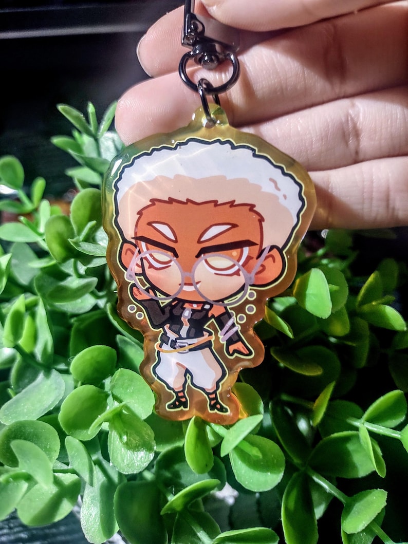 Op de afbeelding: Een heldere sleutelhanger in cartoonstijl met een personage met wit haar, een bril en een zwart-witte outfit. De sleutelhanger heeft een gouden rand en een zwarte sluiting. Het personage staat in een dynamische pose, met een bubbeleffect.