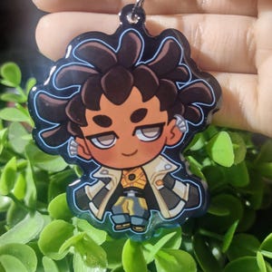 Op de afbeelding: Een sleutelhanger in cartoonstijl met een personage met bruin haar en een geel en blauw kostuum. De sleutelhanger heeft een zwarte rand en een zilverkleurige sluiting. Het personage heeft een bruine huidskleur.