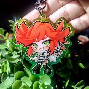 Op de afbeelding: Een kleurrijke anime-stijl sleutelhanger met een personage met rood haar en groene ogen, die een schaar vasthoudt. Het personage draagt een witte jas, een gestreept shirt en een zwarte broek. De sleutelhanger heeft een groene rand en een zilveren sluiting.