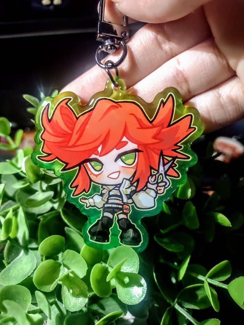 Op de afbeelding: Een kleurrijke anime-stijl sleutelhanger met een personage met rood haar en groene ogen, die een schaar vasthoudt. Het personage draagt een witte jas, een gestreept shirt en een zwarte broek. De sleutelhanger heeft een groene rand en een zilveren sluiting.