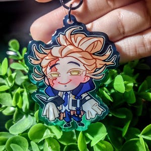 Op de afbeelding: Een kleurrijke anime-stijl sleutelhanger met een personage met blond haar, een blauw-witte outfit en een kleine glimlach. De sleutelhanger heeft een groene rand en een zilveren sluiting.