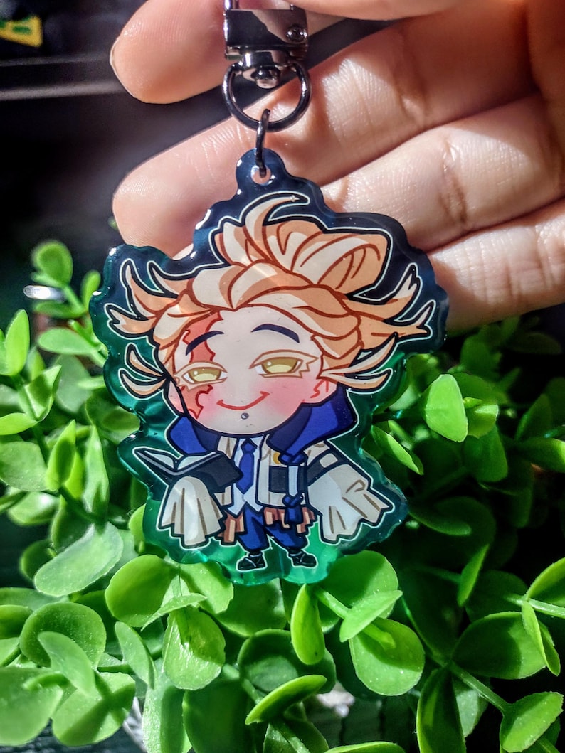 Op de afbeelding: Een kleurrijke anime-stijl sleutelhanger met een personage met blond haar, een blauw-witte outfit en een kleine glimlach. De sleutelhanger heeft een groene rand en een zilveren sluiting.