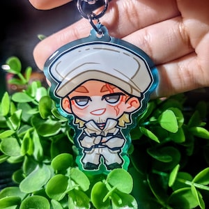 Op de afbeelding: Een heldere acryl sleutelhanger met een cartoon illustratie van een personage met een witte hoed en outfit. De sleutelhanger heeft een zilveren sluiting. Omringd door groen gebladerte.