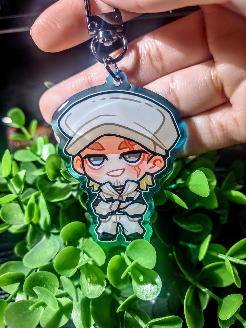Op de afbeelding: Een heldere acryl sleutelhanger met een cartoon illustratie van een personage met een witte hoed en outfit. De sleutelhanger heeft een zilveren sluiting. Omringd door groen gebladerte.