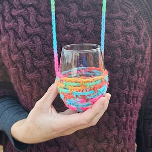 Puede incluir: Un portavasos de vino de crochet colorido con un patrón de arcoíris. El portavasos se lleva como un collar y está hecho de hilo.