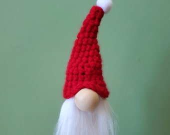 May include: A red crocheted gnome hat with a white pom pom on top. The gnome has a white beard and a wooden nose.